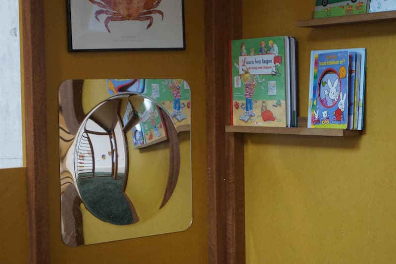 Væg med lille reol med børnebøger og et spej i børnehøjde.